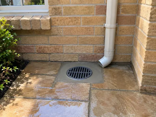 Outdoor drain grate set in wet stone paving beside a brick house wall in Epping